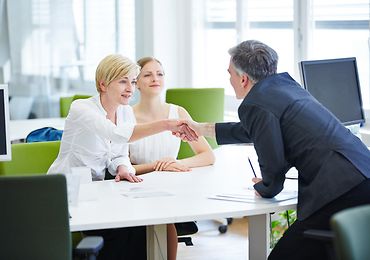 Zwei Frauen und ein Mann sitzen an einem Tisch und schütteln sich die Hände 